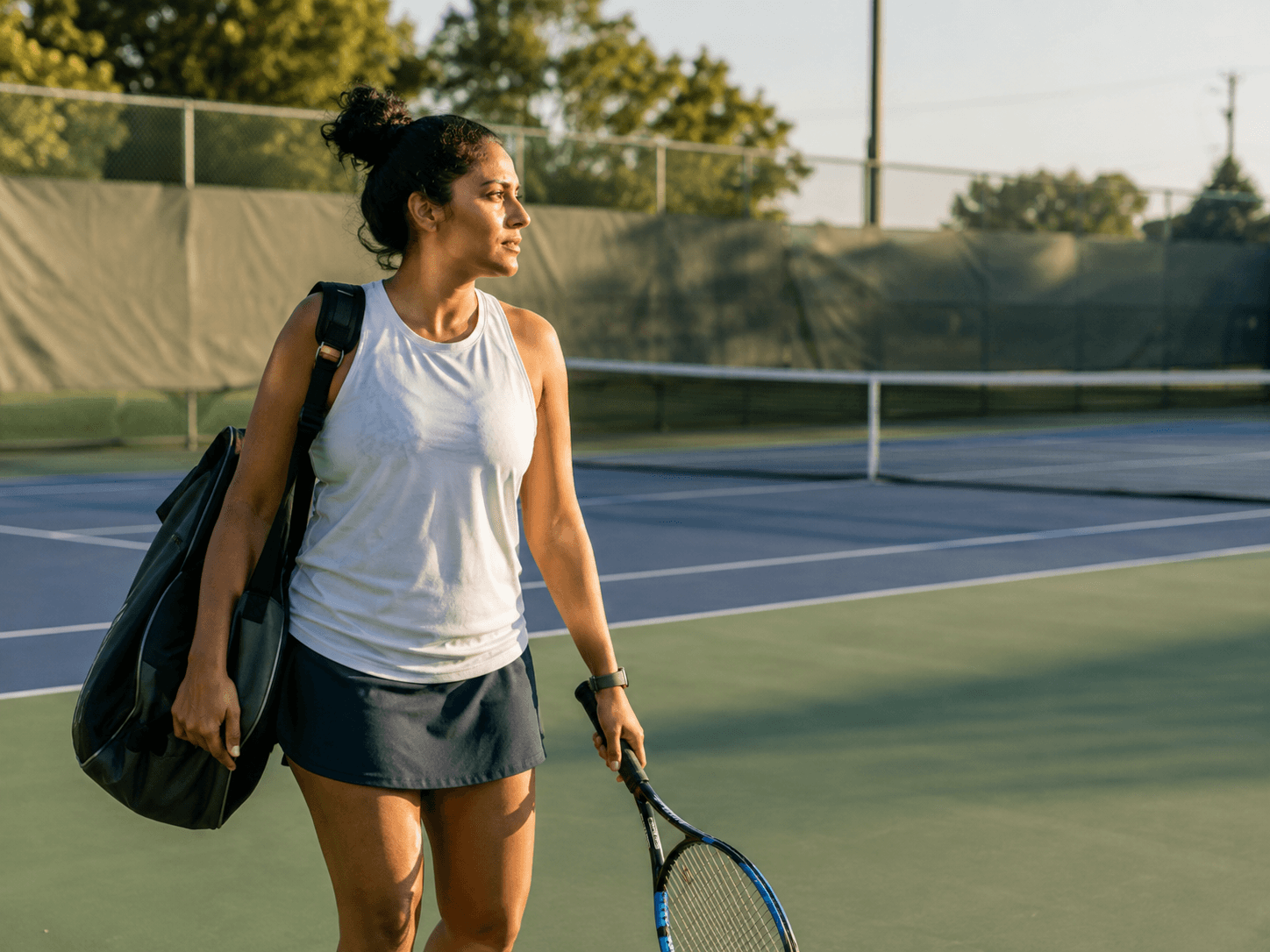 A recreational tennis player at a public court, bag down, picking up a racket before a session.
